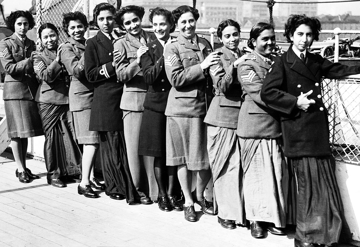 A black and white photo of Indian women in military uniforms posing in a row next to a fence.