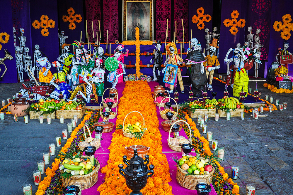 A colorful altar to Our Lady of Guadalupe, heaped with gifts of fruit and flowers and flanked by skeletal figures.