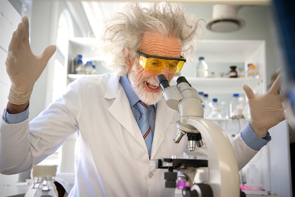 A white man with a shock of white hair looking into a microscope with an excited expression.
