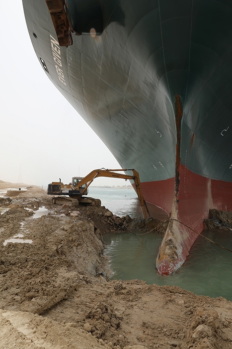 Image of a very small excavator trying to dig out a very large container ship stuck in the mud.